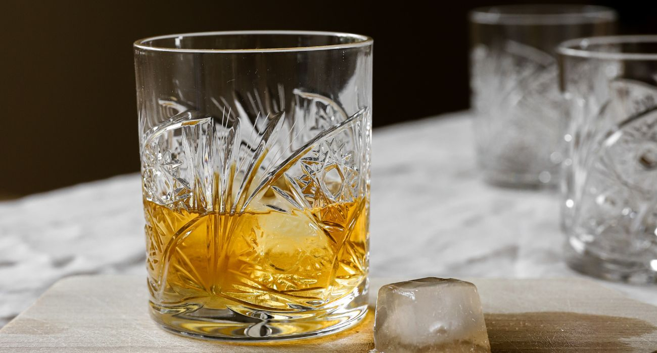 Kryształowa szklanka do whisky z dekoracyjnym szlifem, zawierająca whisky z dużą kostką lodu, ustawiona na drewnianej desce.